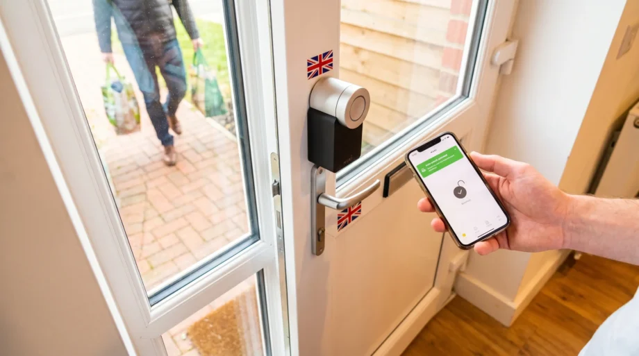 Nuki Smart Lock Pro Review UK — smart lock mounted on inside of white UPVC door with smartphone showing auto-unlock activated, person with shopping bags approaching outside, Union Jack sticker visible